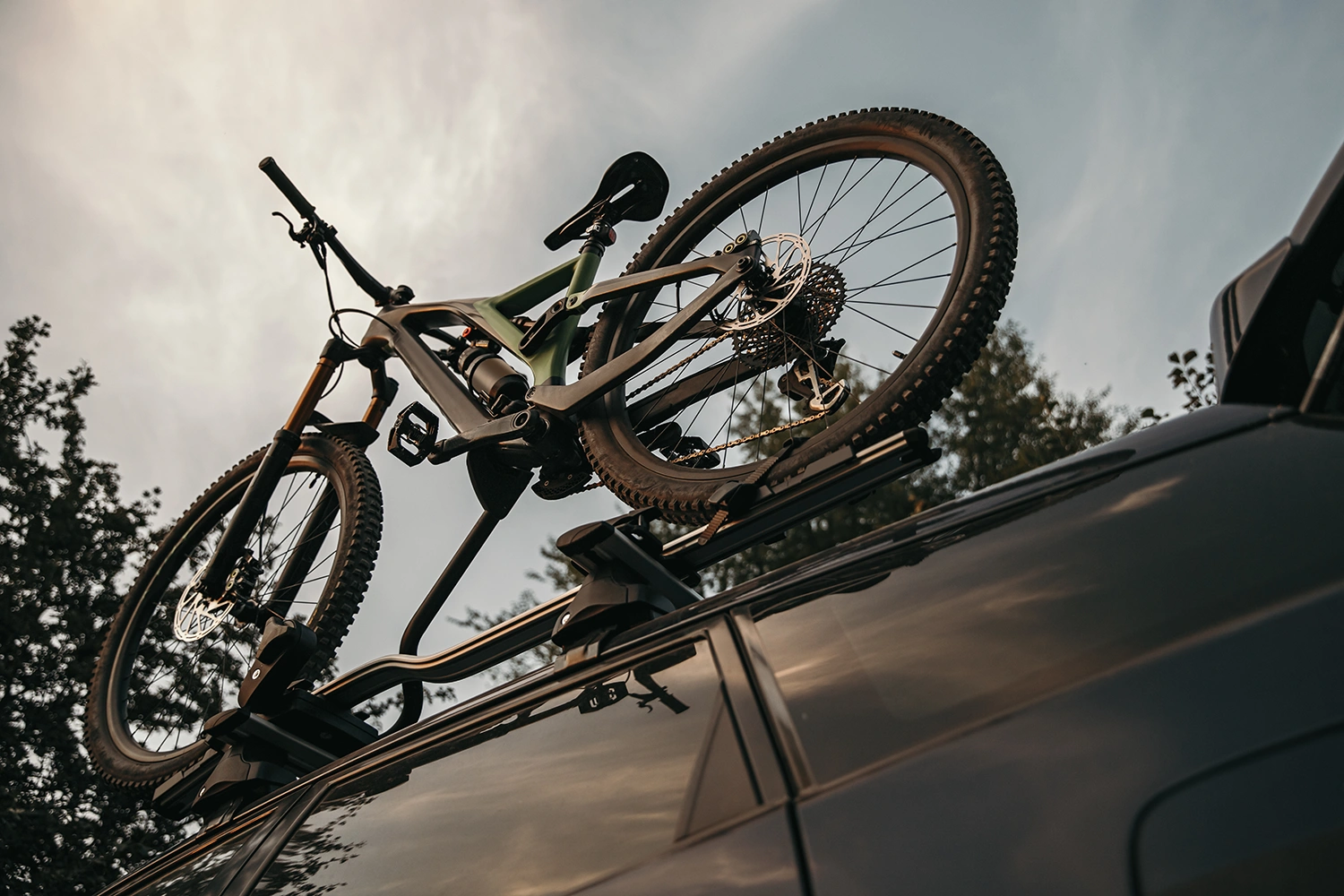 Ein schwarzes Mountainbike, das mit einer Dachhalterung auf dem Dach eines SUVs befestigt ist. Ansicht von unten, Nahaufnahme des Fahrrads. Im Hintergrund sind Bäume und ein bewölkter Himmel zu sehen.