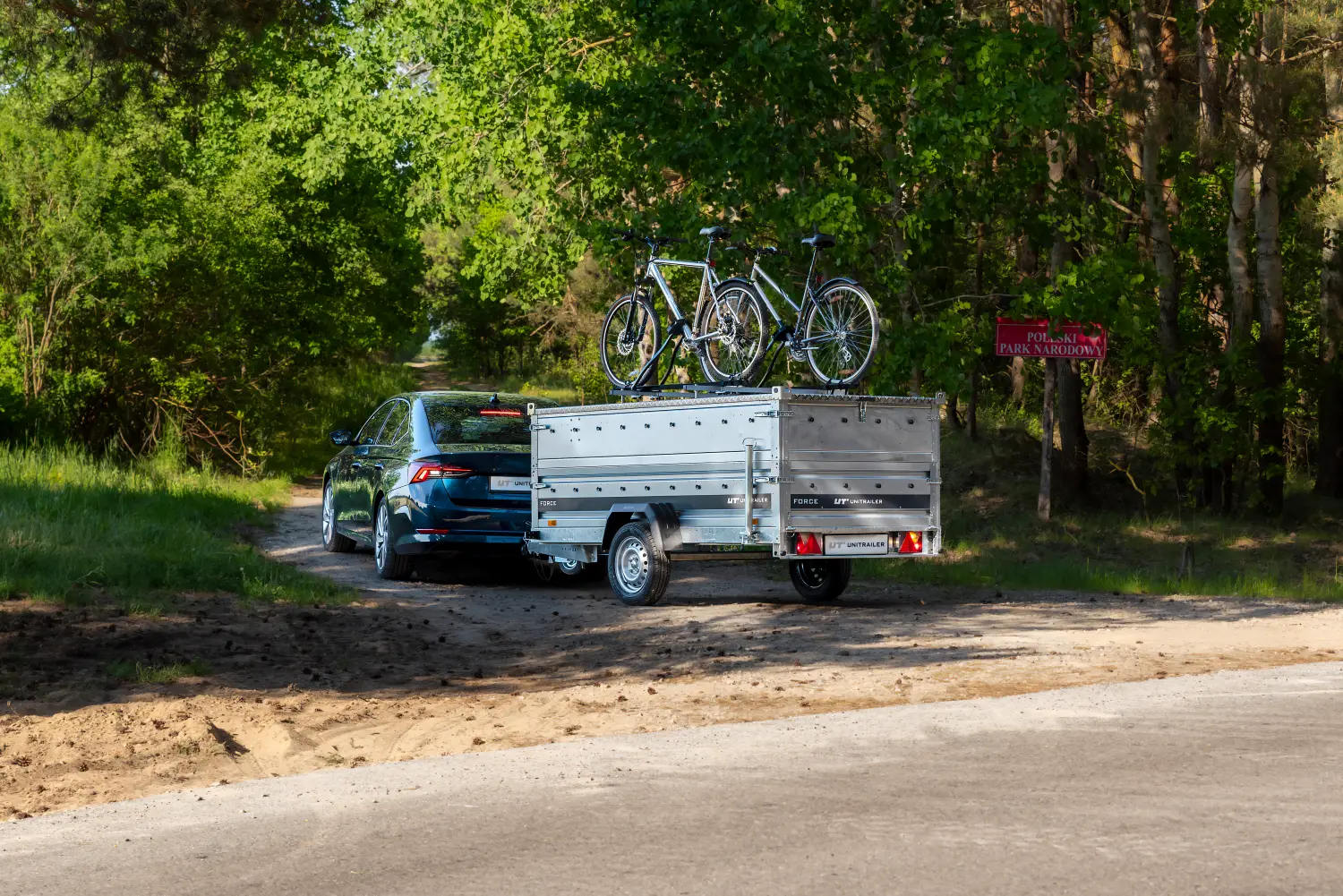 Ein dunkler Kombi mit einer einachsigen Unitrailer-Gepäckanhängerkupplung, ausgestattet mit hohen Bordwänden und zwei Fahrrädern auf der Abdeckung, steht an einer Waldstraße mit einem Schild des Nationalparks.