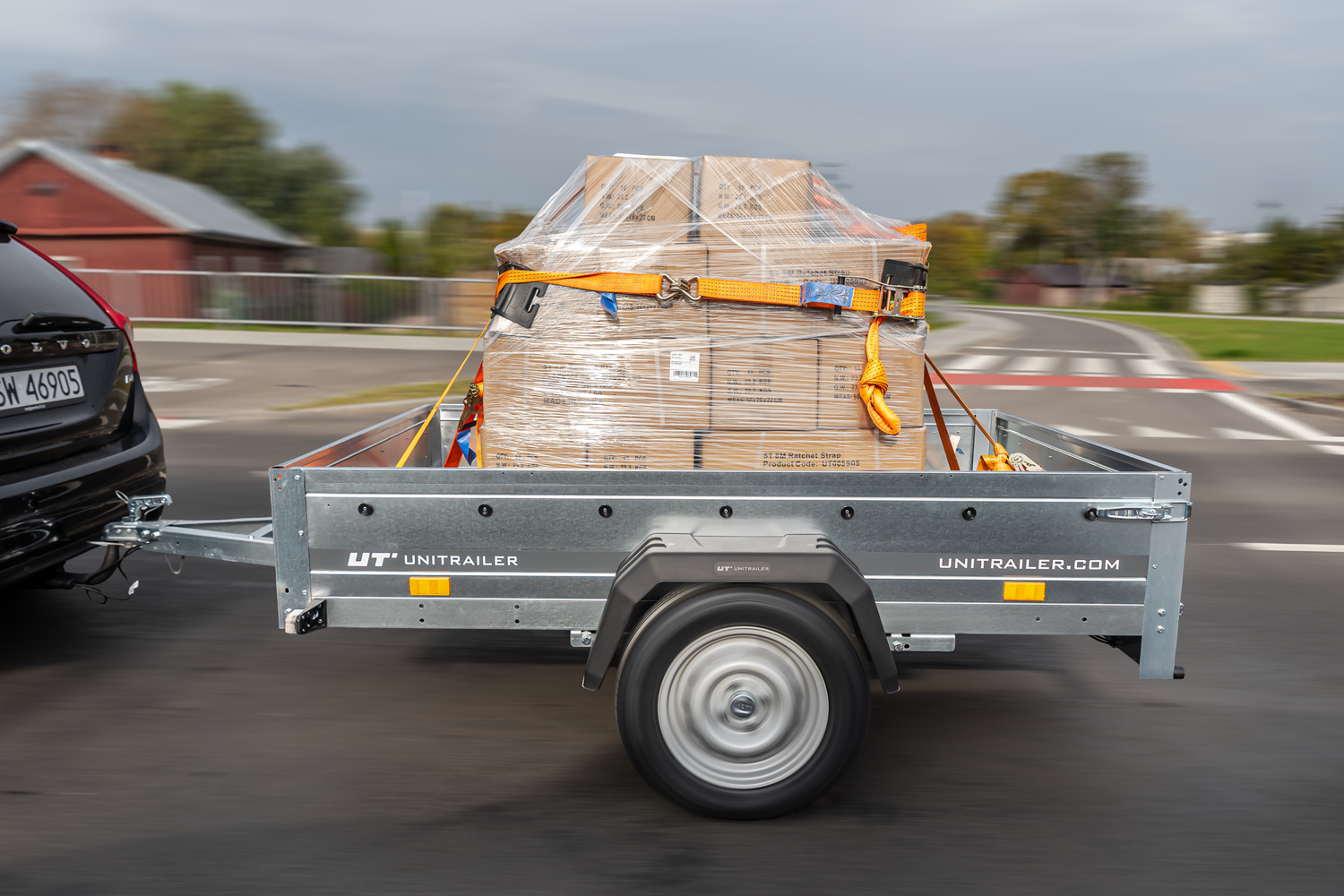 Ein Auto zieht einen Unitrailer-Anhänger mit in Folie gewickelten Kartons, die mit Spanngurten gesichert sind, während der Fahrt auf einer Straße.