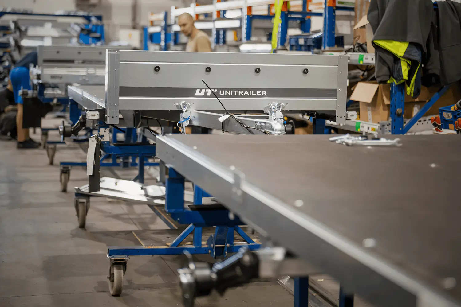 Eine detaillierte Aufnahme aus der Montagehalle, fokussiert auf ein Anhänger-Chassis während des Produktionsprozesses. Mittig im Bild ist die verzinkte, silberne Bordwand eines Anhängers zu sehen, auf der das Markenlogo „UT UNITRAILER“ auf einem dunklen Streifen aufgedruckt ist. Die Konstruktion ruht auf einem blauen, fahrbaren Montagegestell mit Rollen. Der unmittelbare Vordergrund zeigt die angeschnittene Kante einer dunklen Ladefläche, während im verschwommenen Hintergrund ein Fabrikarbeiter und blaue Hochregale zu erkennen sind.