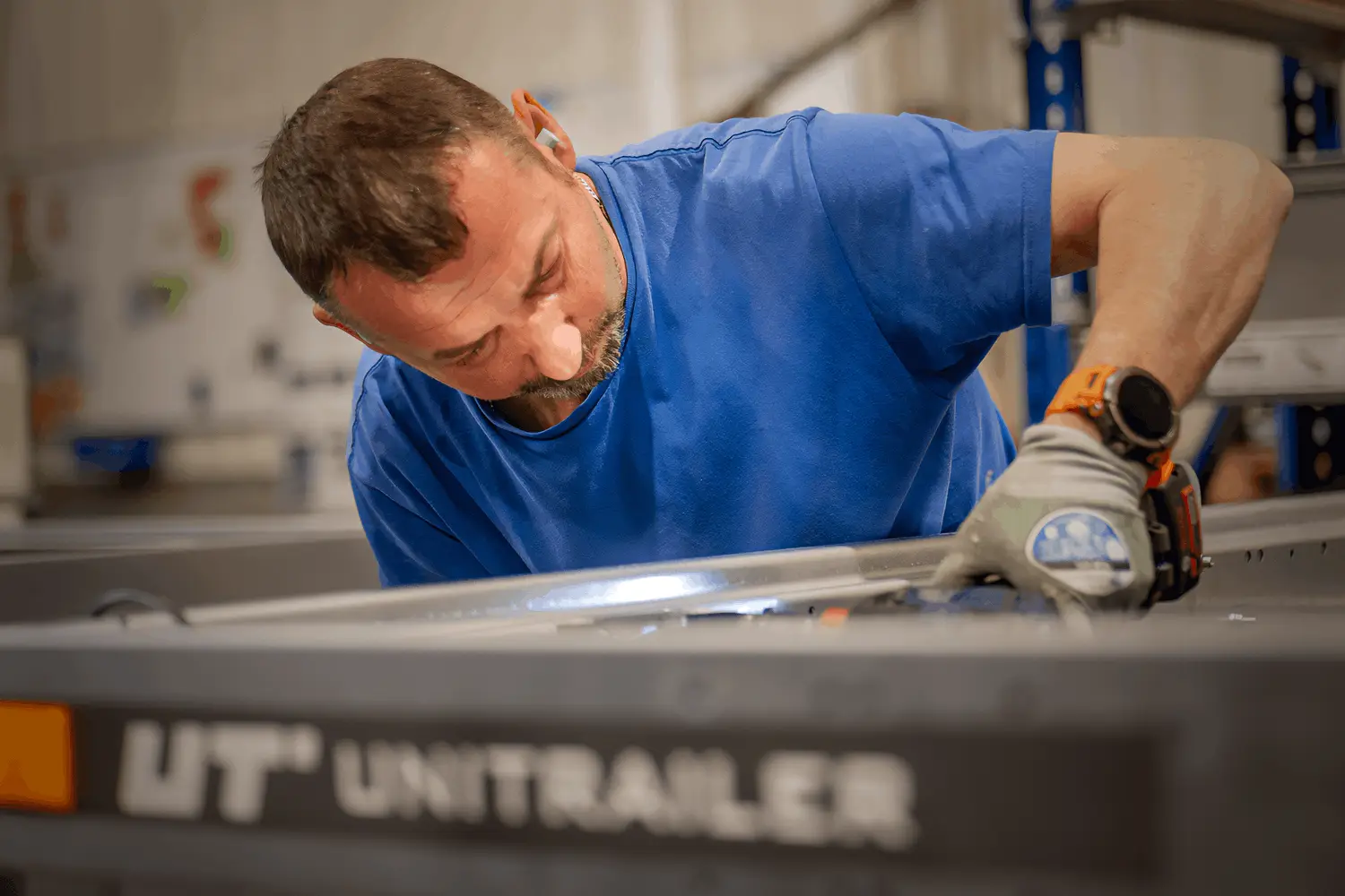 Fokussierte Nahaufnahme eines Handwerkers im blauen T-Shirt und mit hellen Schutzhandschuhen, der konzentriert an der oberen Kante einer Metallkonstruktion arbeitet. Im unscharfen Vordergrund ist der Markenname „UT UNITRAILER“ auf einer grauen Bordwand zu sehen. Der Hintergrund der Werkstatt bleibt dezent in Unschärfe getaucht.