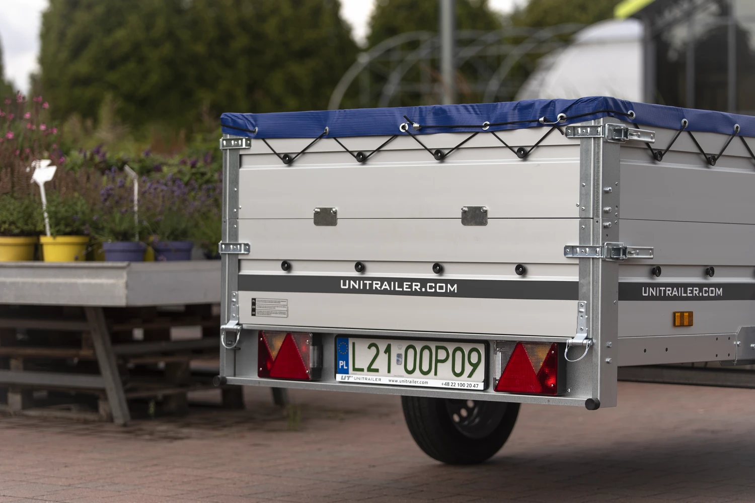 Nahaufnahme der Heckpartie eines Unitrailer-Pkw-Anhängers mit silbernen Aluminium-Bordwänden und gespannter blauer Flachplane. Auf der Heckklappe befindet sich ein schwarzer Streifen mit dem Schriftzug UNITRAILER.COM sowie metallene Verschlusshebel und schwarze Knöpfe zur Planenbefestigung. Im unteren Bereich sind das Kennzeichen L21 00P09, zwei rote Dreiecksrückstrahler und die Rückleuchten zu sehen. Der Anhänger steht auf gepflastertem Boden vor verschwommenen Gartenpflanzen.