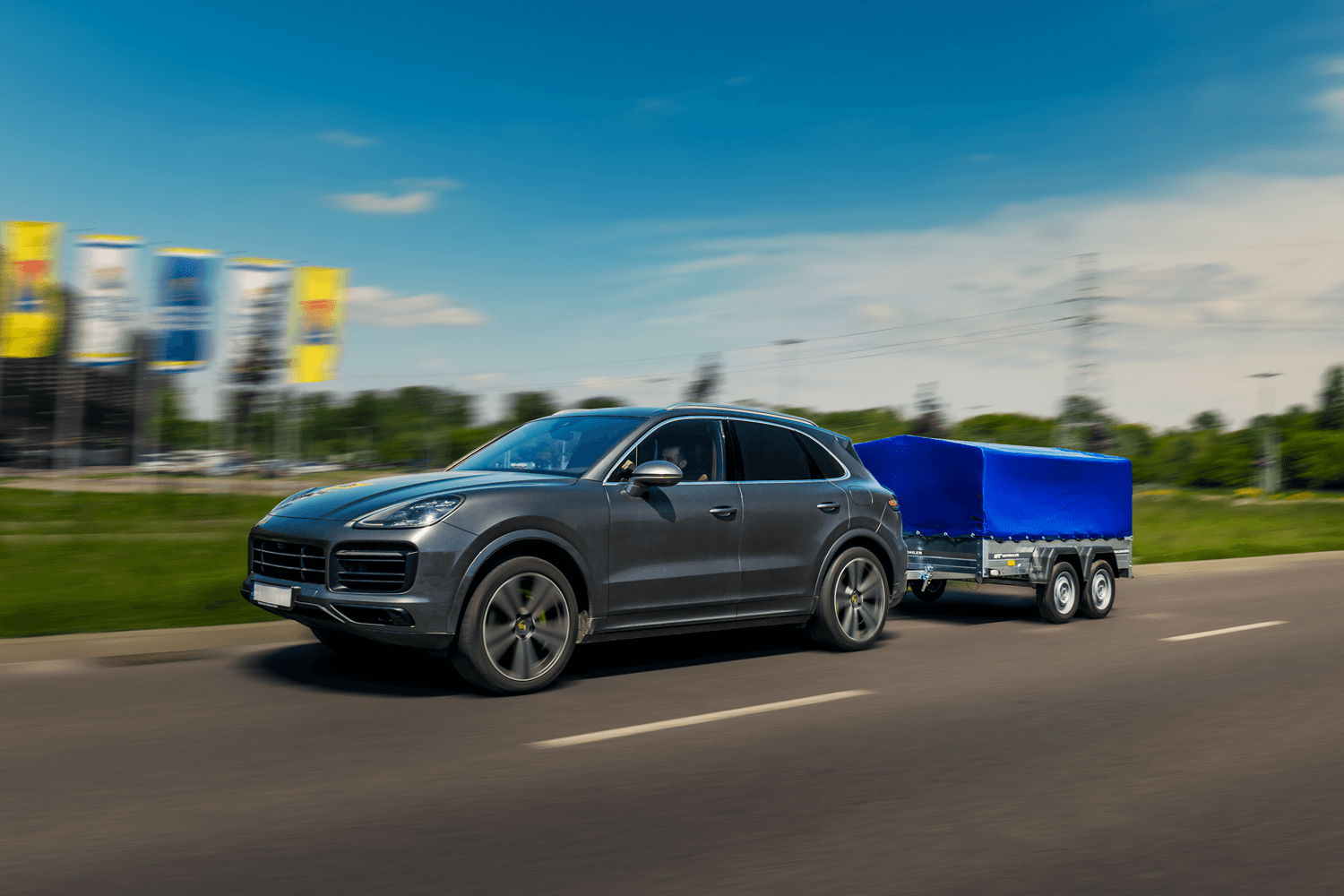 Grauer Porsche Cayenne in Fahrt mit einem zweiachsigen Unitrailer Pkw-Anhänger inklusive blauer Abdeckplane.