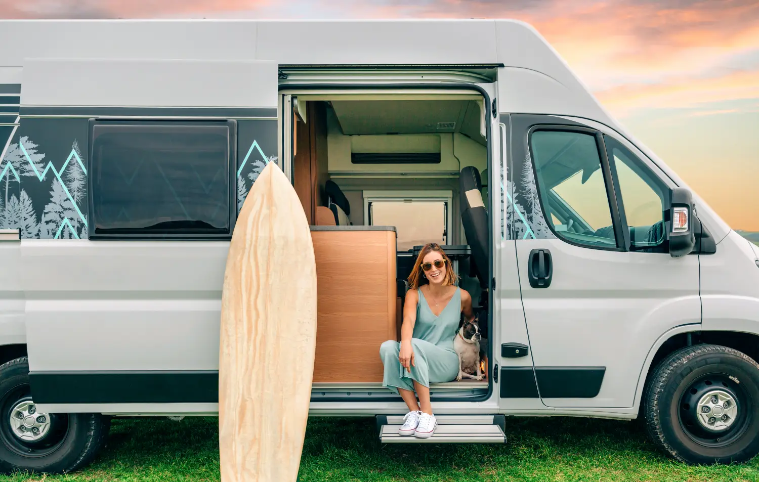 Lächelnde Frau mit Sonnenbrille sitzt mit ihrem kleinen Hund im Eingang eines grauen Campervans mit Berg-Motiv. Neben der geöffneten Schiebetür lehnt ein Surfbrett aus Holz vor einem farbenfrohen Abendhimmel.